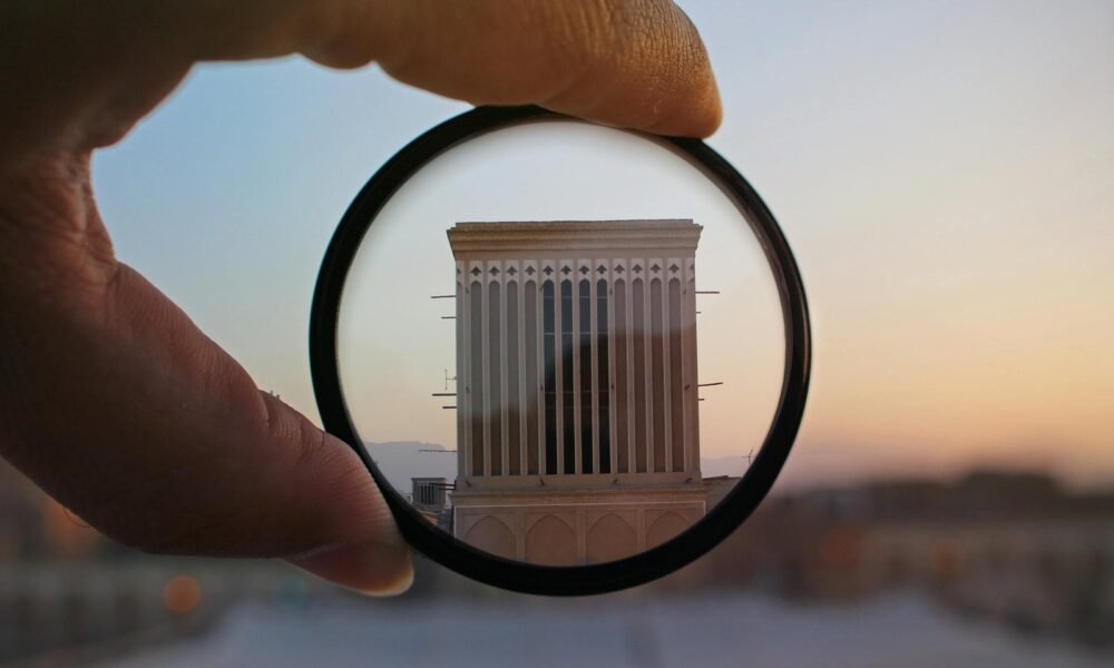 A hand holding a lens magnifies a building facade at sunset, highlighting architectural details in an abstract composition.