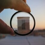 A hand holding a lens magnifies a building facade at sunset, highlighting architectural details in an abstract composition.