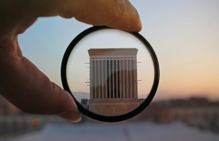 A hand holding a lens magnifies a building facade at sunset, highlighting architectural details in an abstract composition.