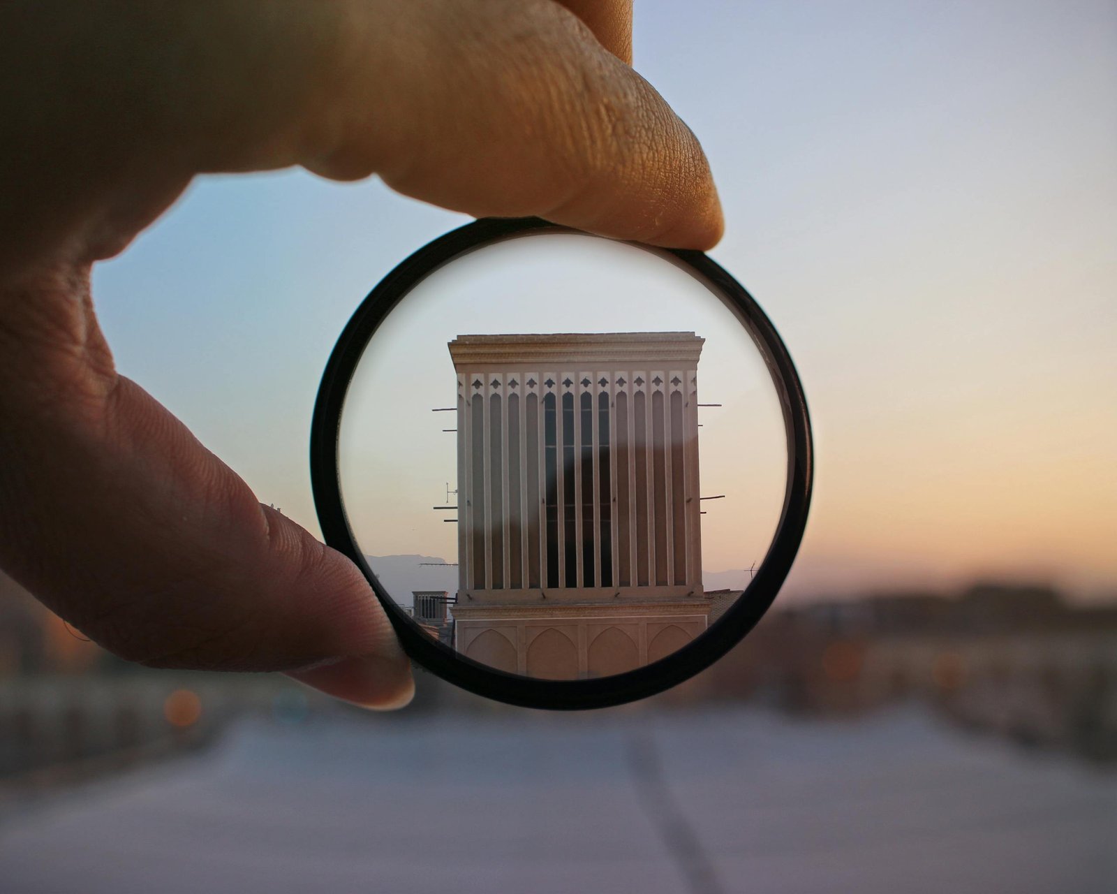 A hand holding a lens magnifies a building facade at sunset, highlighting architectural details in an abstract composition.